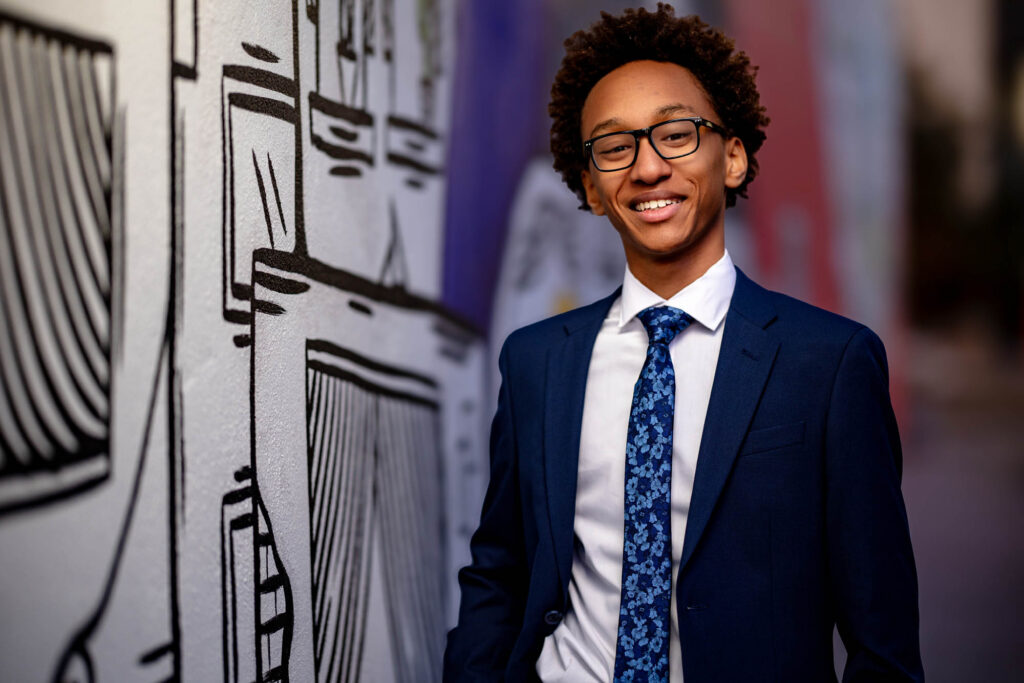 High school senior in navy blazer and blue floral tie laughing beside a graphic mural in downtown La Crosse, WI