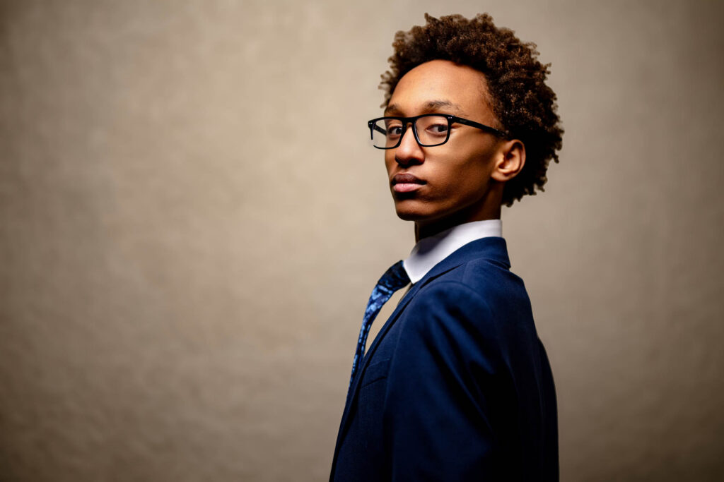 High school senior in navy blazer and blue tie looking directly at camera over his shoulder against a warm concrete wall