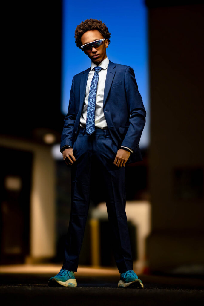 Full-length portrait of a high school senior in a blue suit between buildings with a deep blue sky overhead