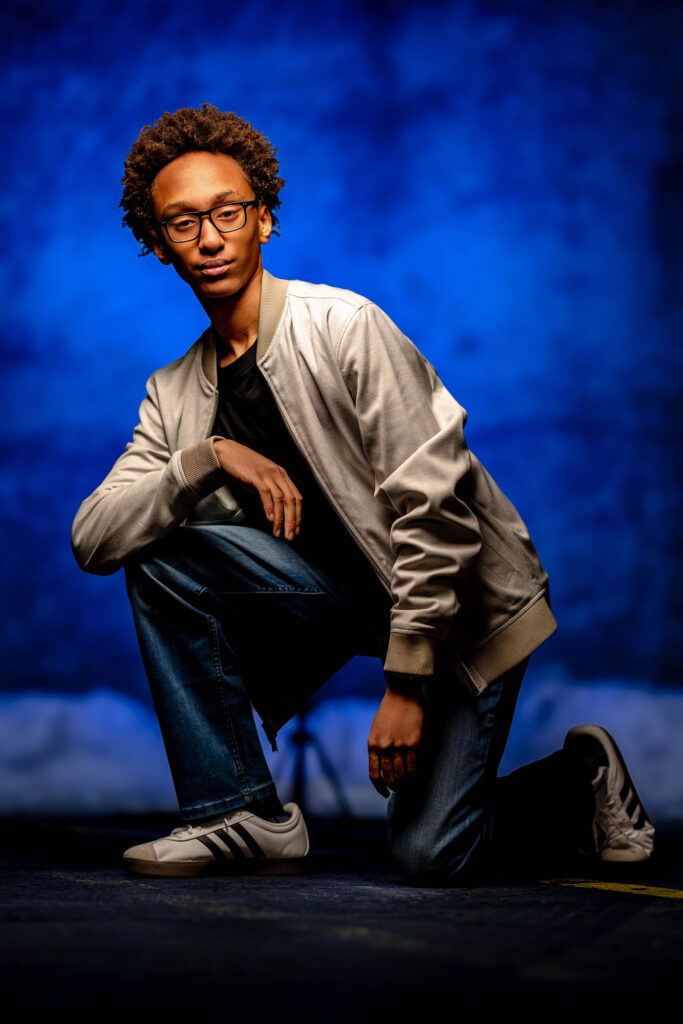 High school senior kneeling on a dark floor with rich blue studio light behind him, gaze direct and composed