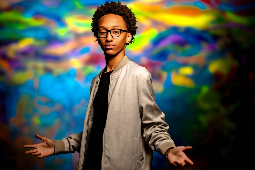 High school senior in grey bomber jacket arms spread wide, intense gaze against a vivid swirling neon backdrop
