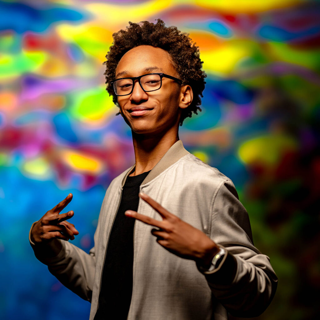 Teen male senior with glasses smiling genuinely while posing with hand signs before a bold multicolored mural