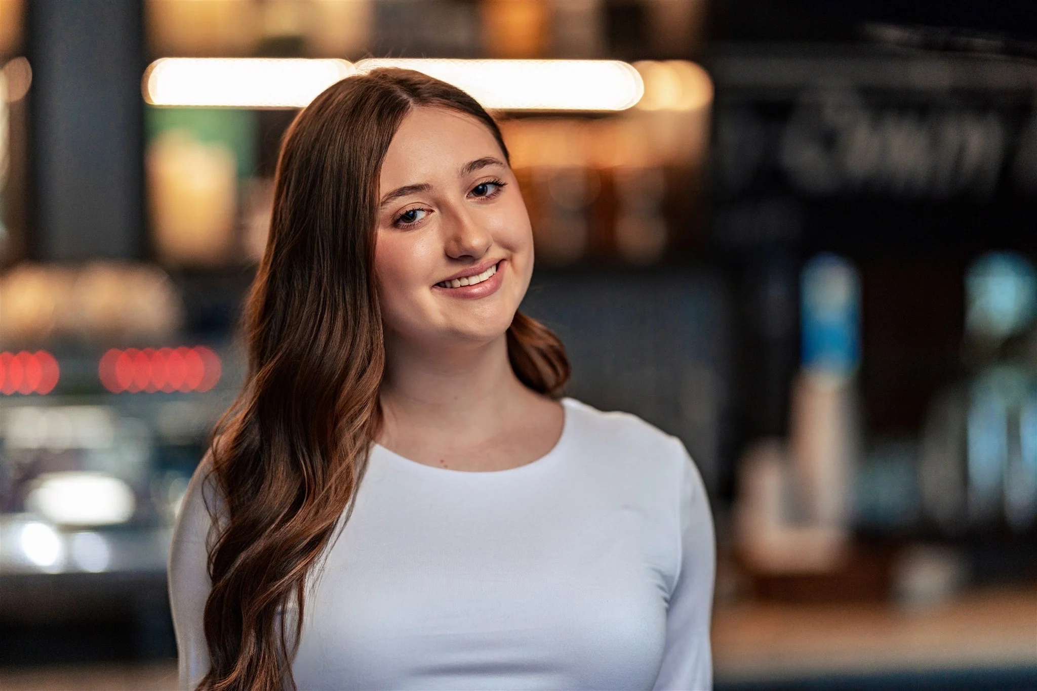 High school senior girl with long brown hair smiling in a white top, photographed at a cafe in La Crosse, Wisconsin.