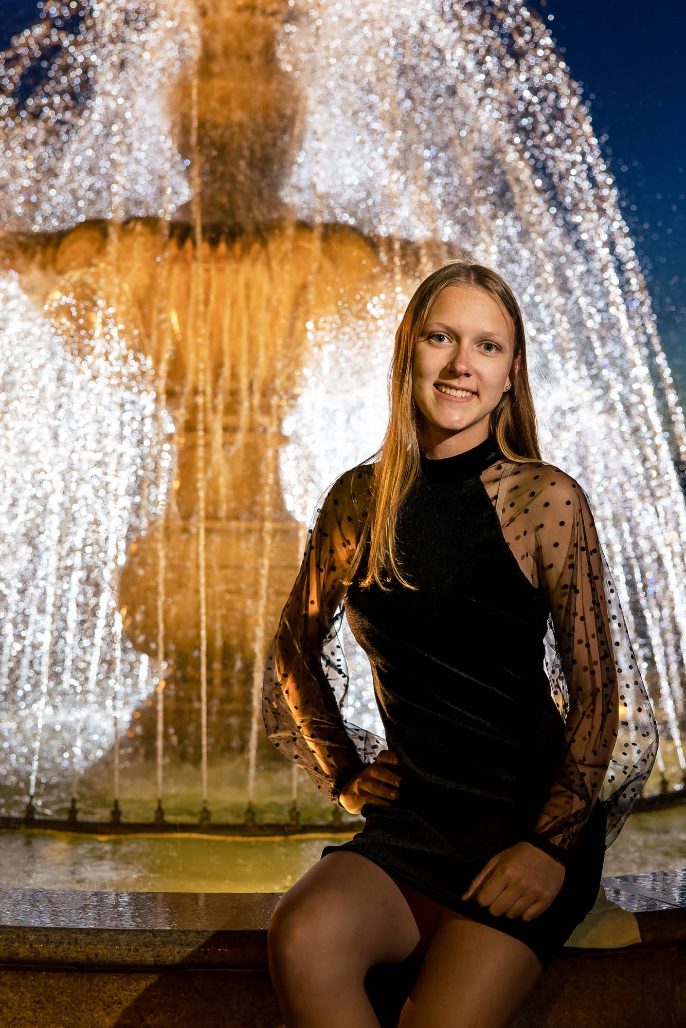 High school senior in a black dress seated in front of a lit fountain at night, La Crosse