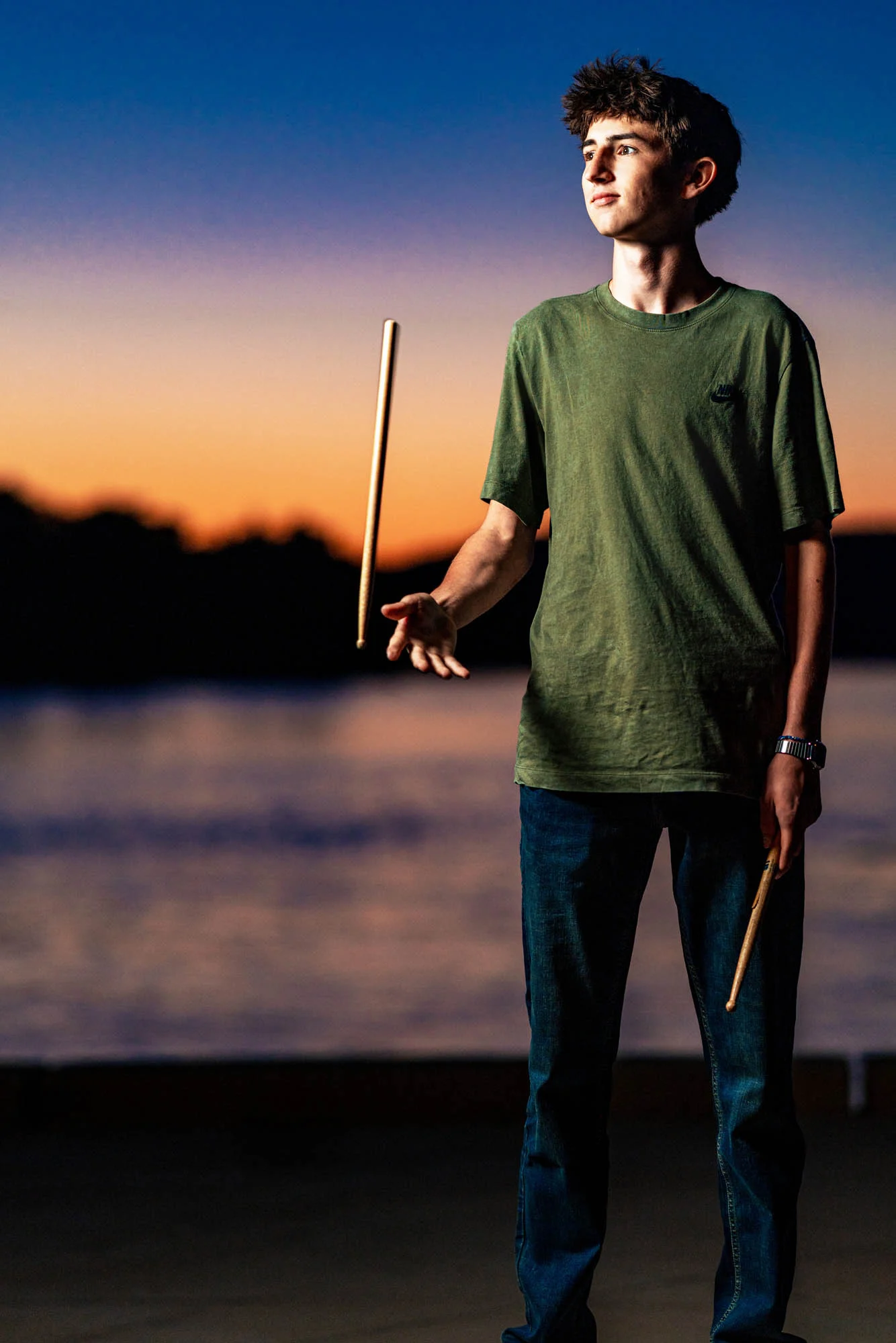 High school senior tossing a drumstick at sunset near the water, La Crosse, lit with flash against vivid dusk sky.