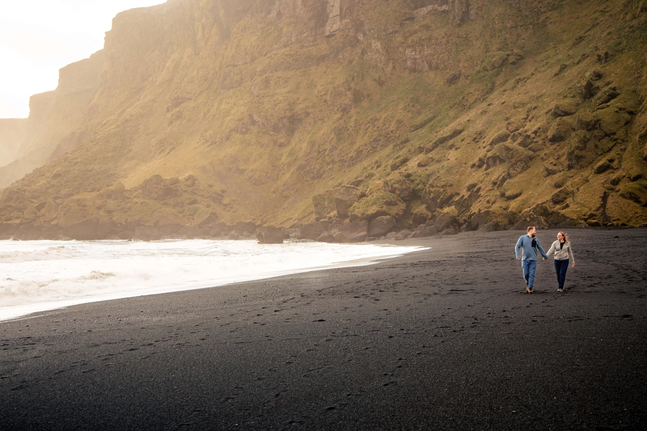 Couple walking hand in hand along a black sand beach beneath green cliffs