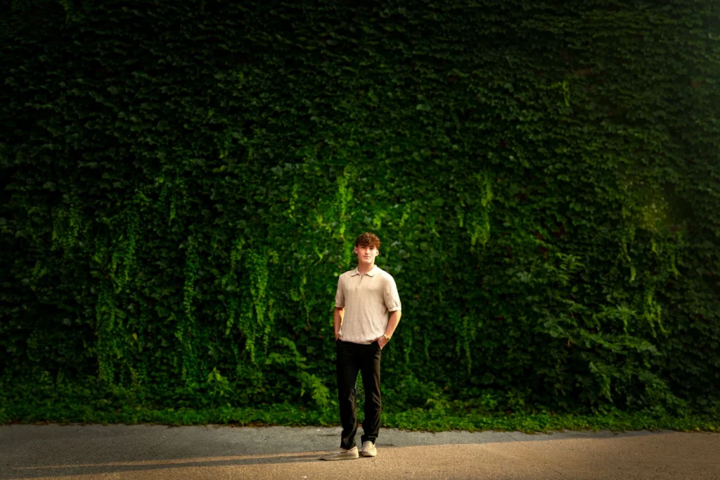 High school senior in cream polo standing before a tall ivy-covered wall in La Crosse Wisconsin