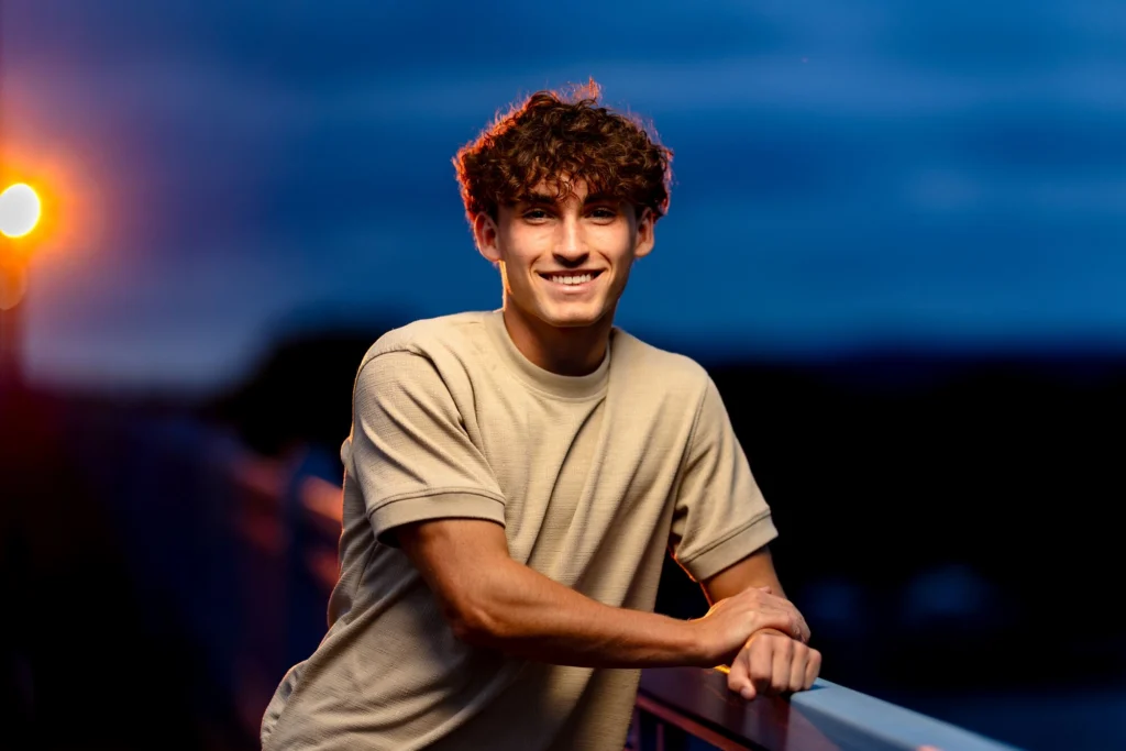 High school senior with curly hair beaming at camera on Cass Street Bridge at dusk with glowing lamplight, La Crosse WI