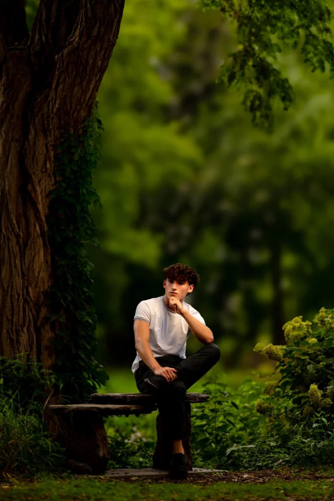 High school senior guy seated on a wooden bench under a large tree in a lush green park, chin resting on hand