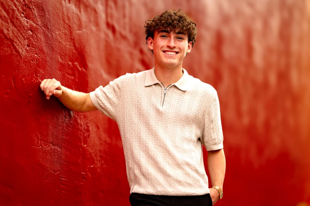 Senior smiling and leaning against a red wall in cream polo shirt, La Crosse Wisconsin senior portrait