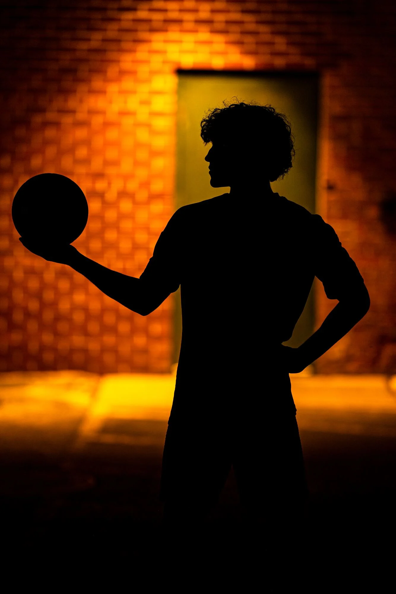 Senior portrait: Deacon in silhouette with a soccer ball against a glowing brick wall, by J.L. Wiswell Photography