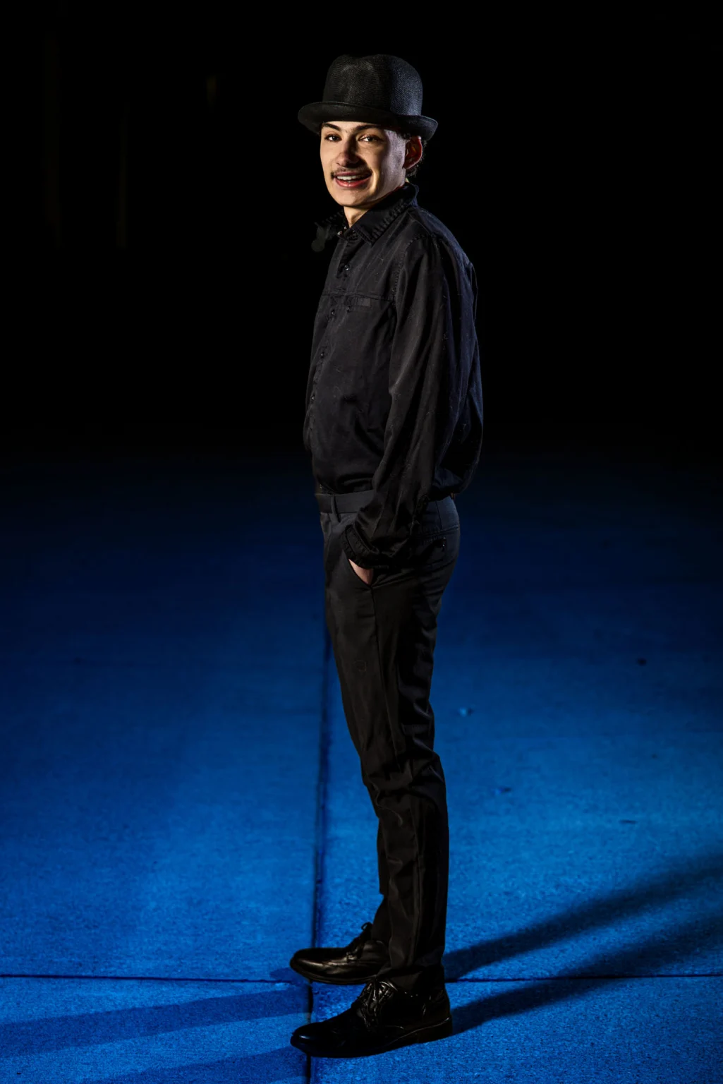 Senior in a black fedora and all-black outfit under dramatic light on a blue floor