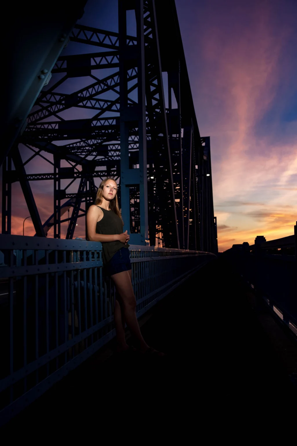 High school senior girl leaning on the Cass Street Bridge in La Crosse at sunset, lit by flash against a purple sky