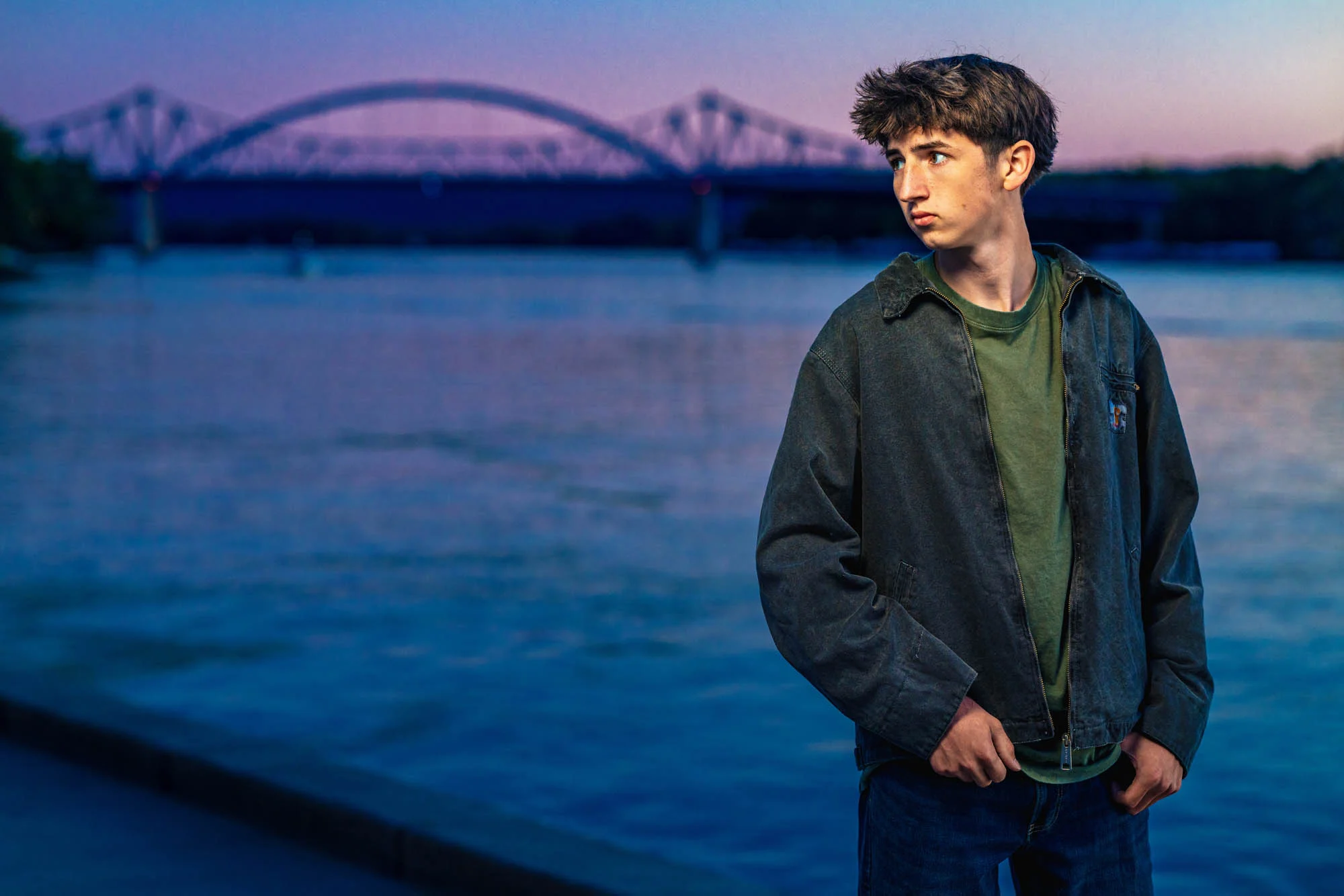 High school senior boy in jacket standing along the Mississippi River at dusk, Cass Street Bridge glowing behind him, La Crosse WI.